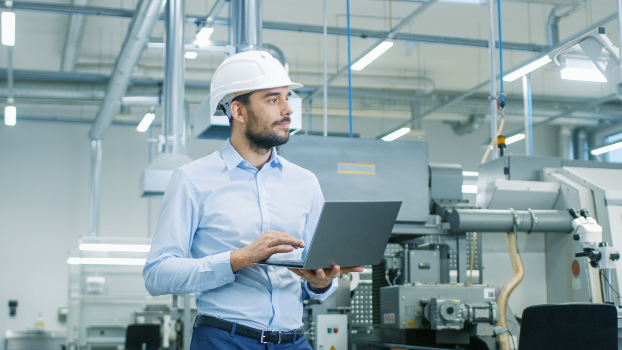 Chief engineer in a hard hat walks through light Modern factory while holding laptop. Successful, Handsome Man in Modern Industrial Environment to represent use of technology in manufacturer rep industry 